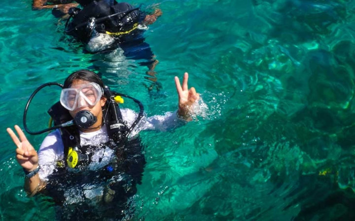A person wearing Scuba gear floats in clear blue-green water and gives the camera two peace signs with both their hands